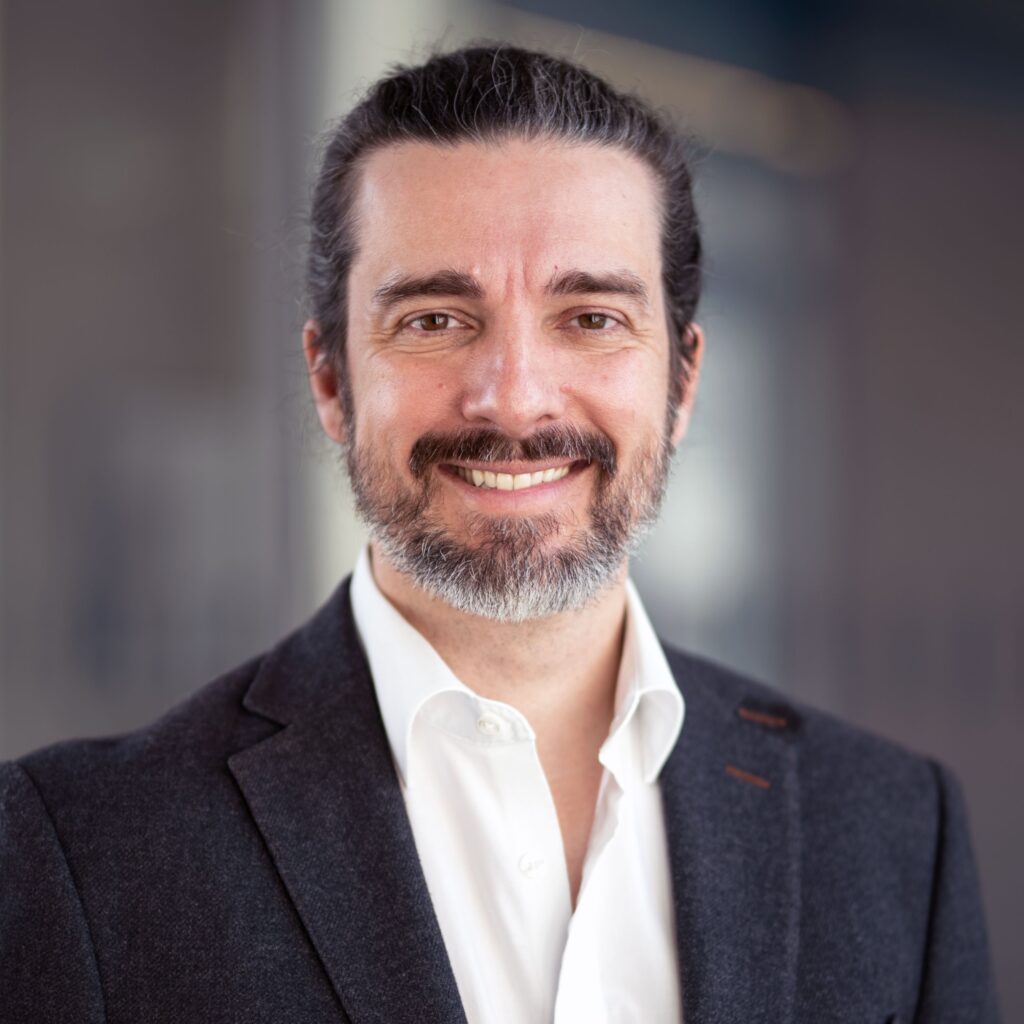 Das Bild zeigt Prof. Dr. Saulo Henrique Freitas Seabra da Rocha. Einen Mann mit dunklen langen Haaren die zurückgebunden sind. Er hat einen dunklen kurzen Vollbart der langsam ergraut. Er trägt ein weißes Hemd und einen Schwarzen Anzug. Er ist von Kopf bis zur Brust zu sehen. 